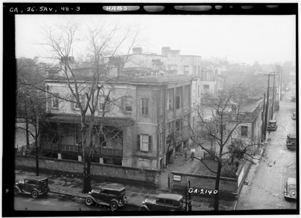 Historic B&W aerial view of a three story home surrounded by a brick wall. The surrounding trees are bare and the streets are wet as if it rained recently. Old Model T Ford-style vehicles are parked in the road out front