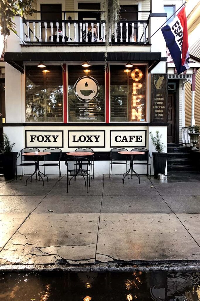 Exterior shot of the front 2-story façade of Foxy Loxy Café in the Starland District. The restaurant is set in a historic home that is painted white with a white balcony on the second level. There are 3 small seating areas along the sidewalk and a red, white, and blue flag with the word "Espresso" hanging above the entrance.