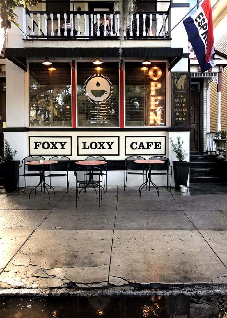 Exterior shot of the front 2-story façade of Foxy Loxy Café in the Starland District. The restaurant is set in a historic home that is painted white with a white balcony on the second level. There are 3 small seating areas along the sidewalk and a red, white, and blue flag with the word "Espresso" hanging above the entrance.