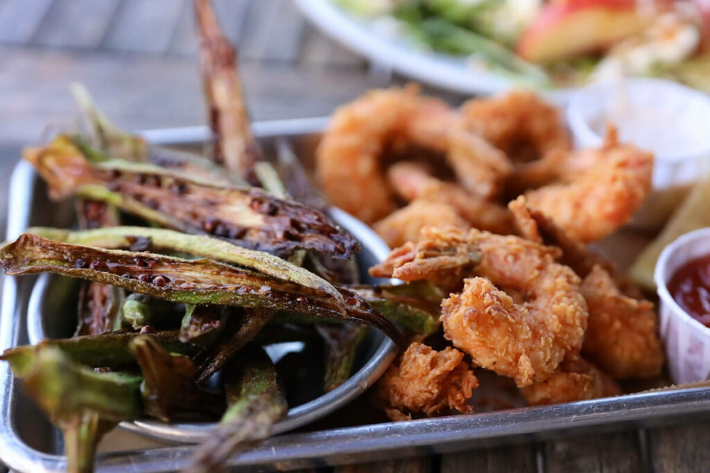 Metal tray loaded with crispy strips of okra and fried shrimp. A salad is visible in the background