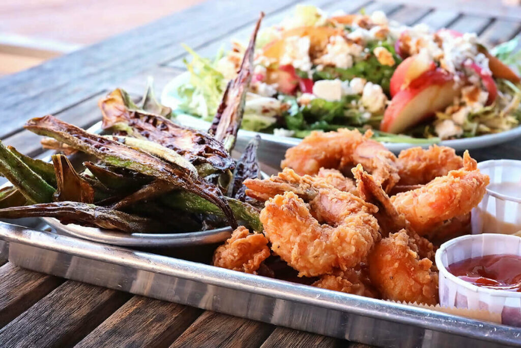 A slatted table made from driftwood with a metal tray full of golden fried shrimp and crispy fried okra. A colorful salad is visible in the background