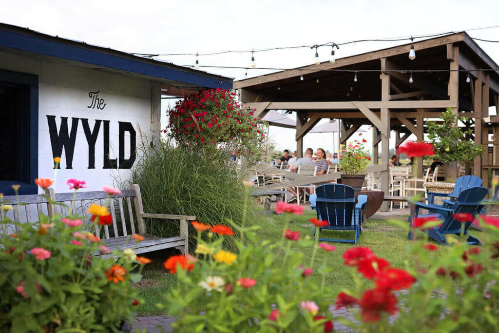 Colorful landscaping at The Wyld, a dog-friendly restaurant in Savannah. Red, orange, and yellow poppies are visible in the foreground, while blue Adirondack chairs surround a fire pit and a covered eating area is visible in the background. Stringlights hang above the scene