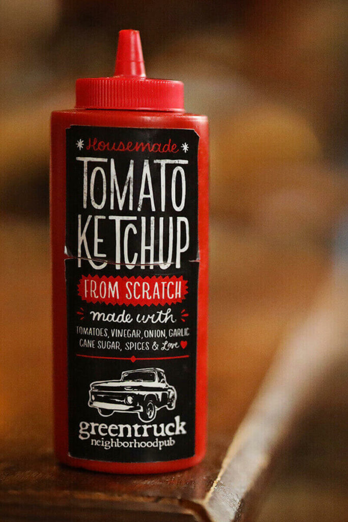 Close-up shot of a red bottle of ketchup with a B&W label that says "Housemade Tomato Ketchup From Scratch - Made with tomatoes, vinegar, onion, garlic, cane sugar, spices, and LOVE". The Green Truck Pub logo with an old-timey truck is visible at the bottom