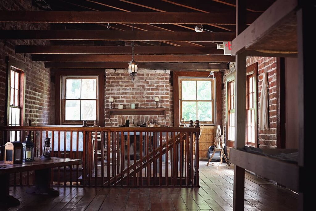 Two simple rooms in the carriage house of the Sorrel Weed House show dark-stained wood floors and trim, simple wooden bunk beds, exposed rafters, exposed brick walls, and a fireplace.