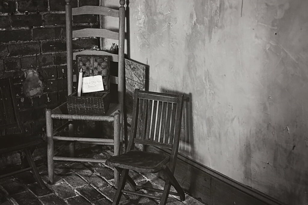 B&W image of three stiff wooden chairs sitting in the corner of a basement that has uneven brick floors and walls