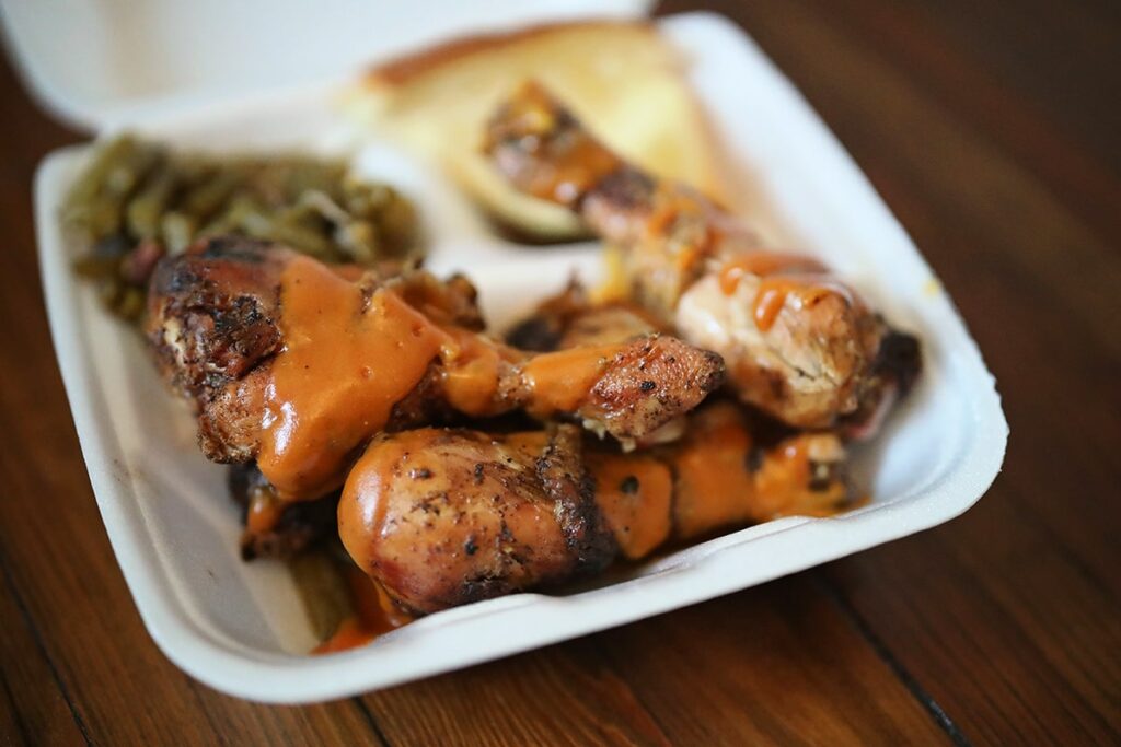 Styrofoam to-go container filled with four plump pieces of BBQ chicken from Munchies BBQ with Carolina Gold BBQ sauce drizzled over the top. Green beens and bread are visible in the background