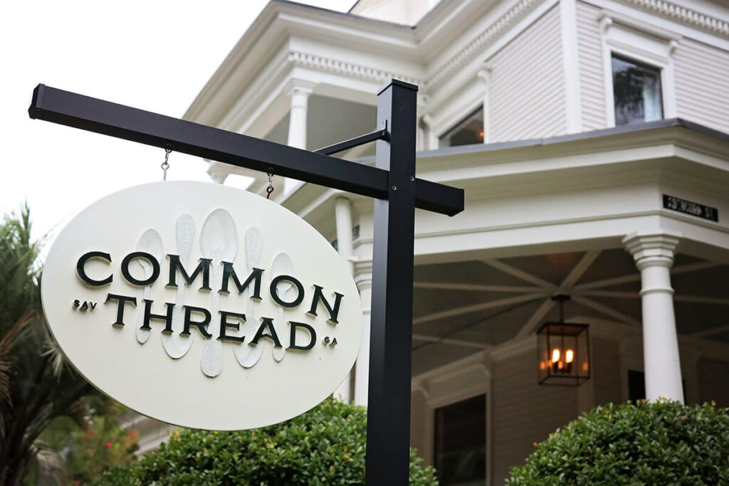 Oval sign with a white background and raised spoon shapes with the words "Common Thread | SAV GA" in black atop of the spoons. In the background a beautiful 3-story home with a large front porch and gas lanterns is visible