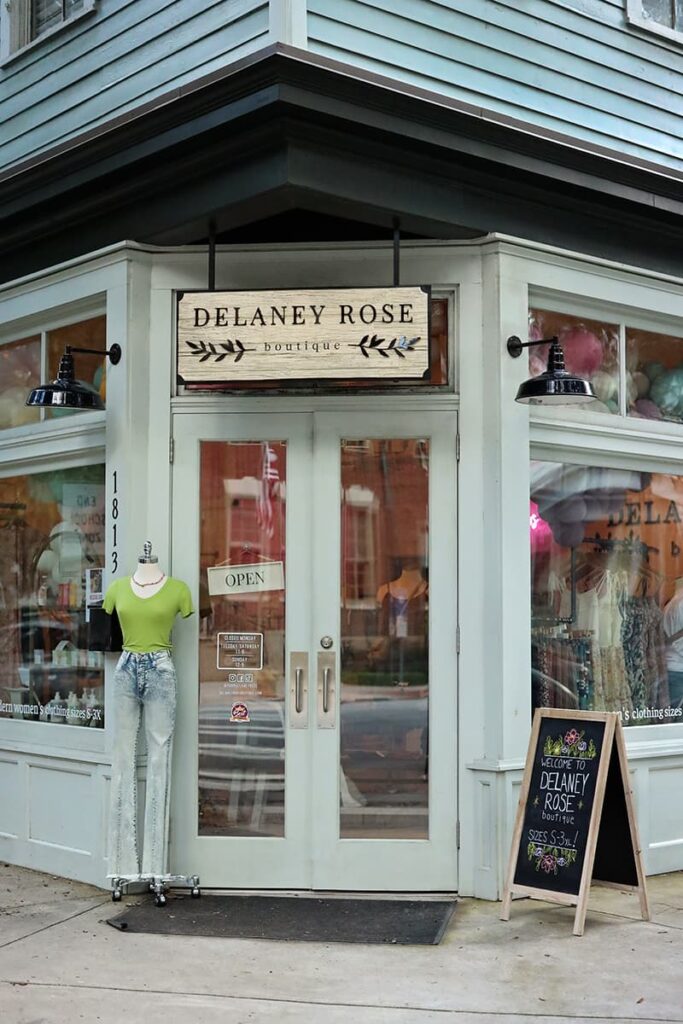 The front entrance to Delaney Rose Boutique in the Starland District with double glass doors and windows allowing a sneak peek into the shop. All of the trim is painted a pale, sage green, and a wooden sign with the store's logo painted in black hangs above the entrance
