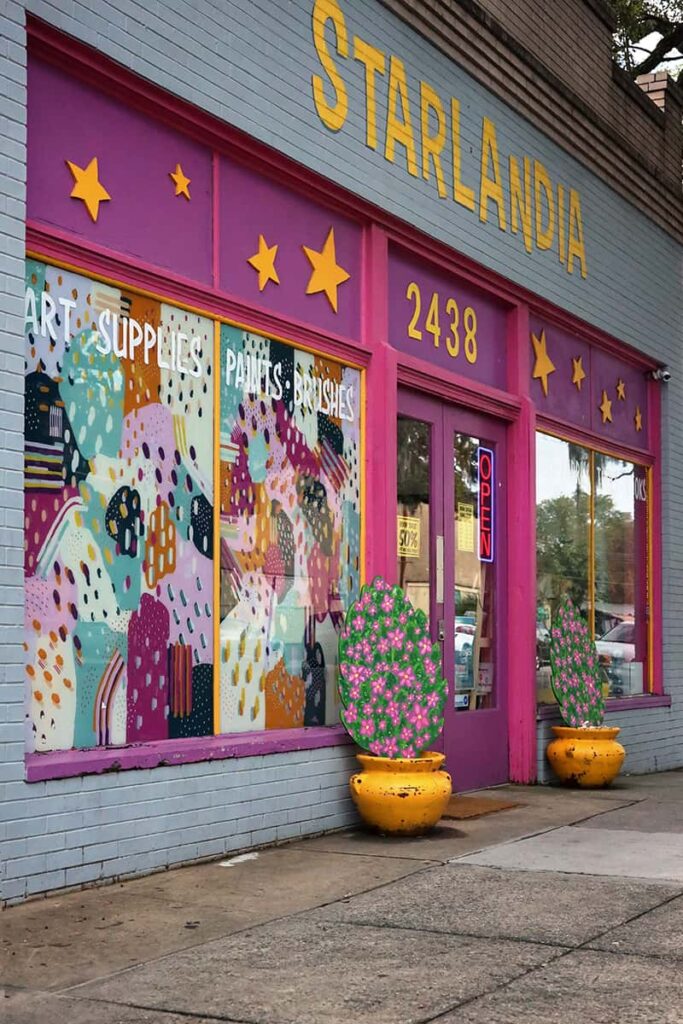 Front view of the Starlandia shop in Savannah's Starland District with grey-painted bricks, hot pink paint, and bright yellow star accents. One window has a colorful mural and the sign reads "Art Supplies | Paints | Brushes"