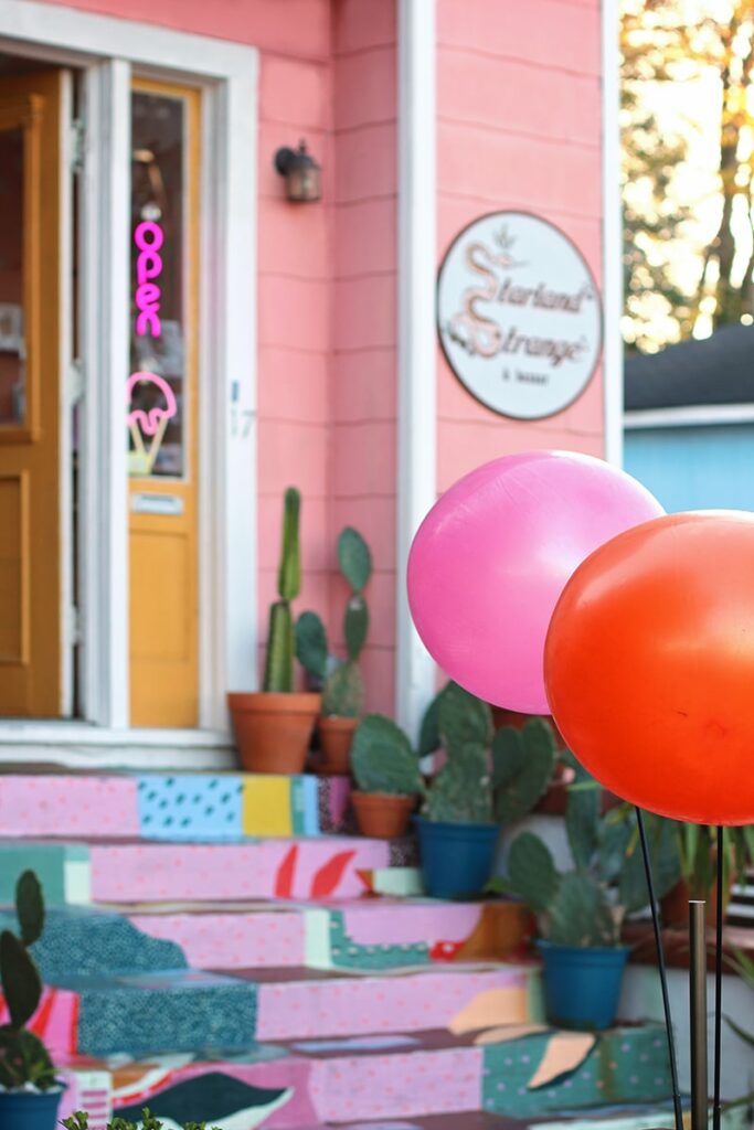 The colorful front entrance to Starland Strange, one of the quirkier Starland District shopping options, shows a pink wall with white trim and a mustard-colored front door. Hot pink and red balloons line the front walk, which is covered by a colorful mural