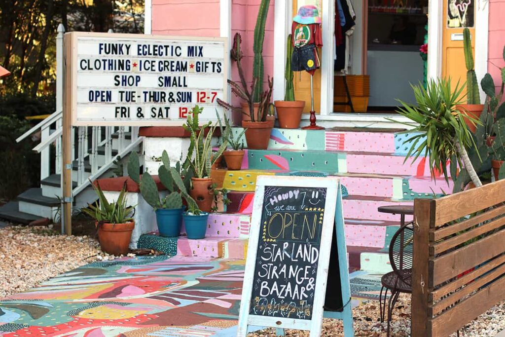 The colorful front entrance to Starland Strange & Bazaar shows a sidewalk and steps painted with a colorful mural, stairs lined with cacti, and a sign indicating the stores hours and "funky eclectic mix" of items