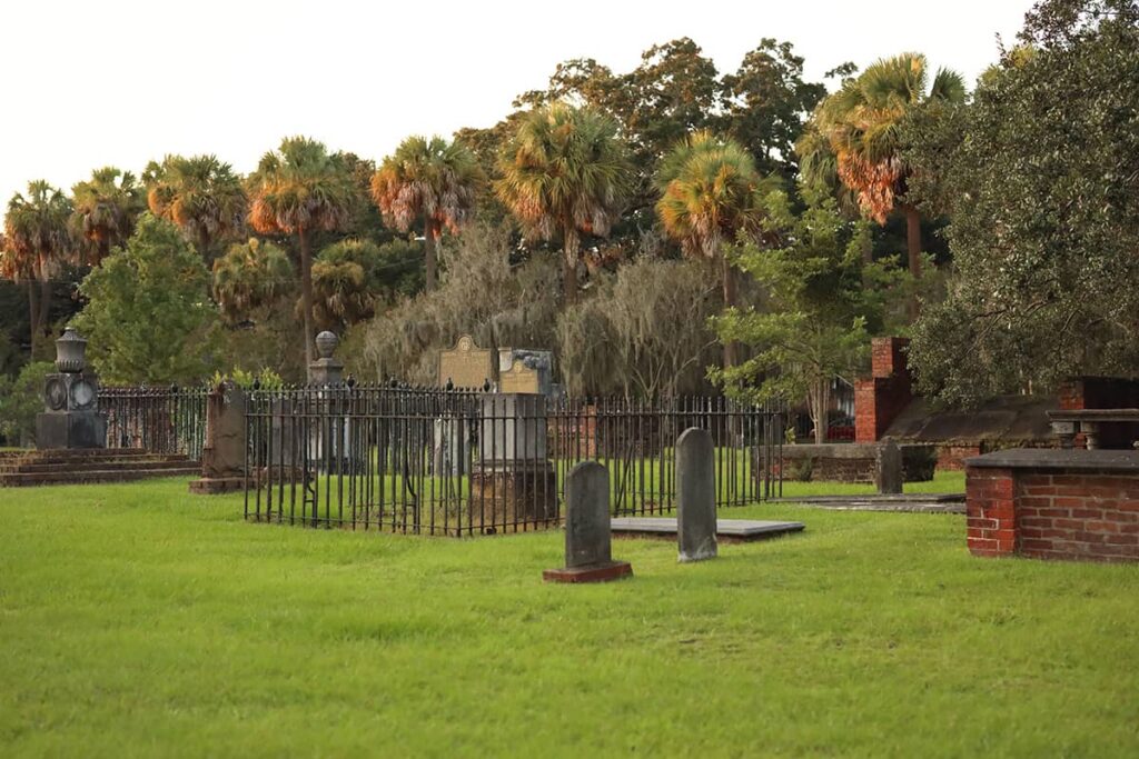 A wide angle of numerous headstones and burial plots in Colonial Park Cemetery with fall colors showing in the background as the sunset illuminates a row of palm trees