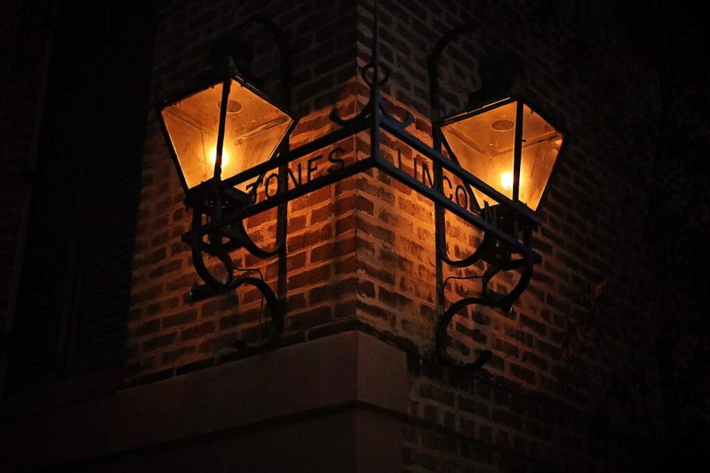 Nighttime scene at the corner of Jones and Lincoln streets in Savannah showing two elaborate gas lantern attached to a historic brick home