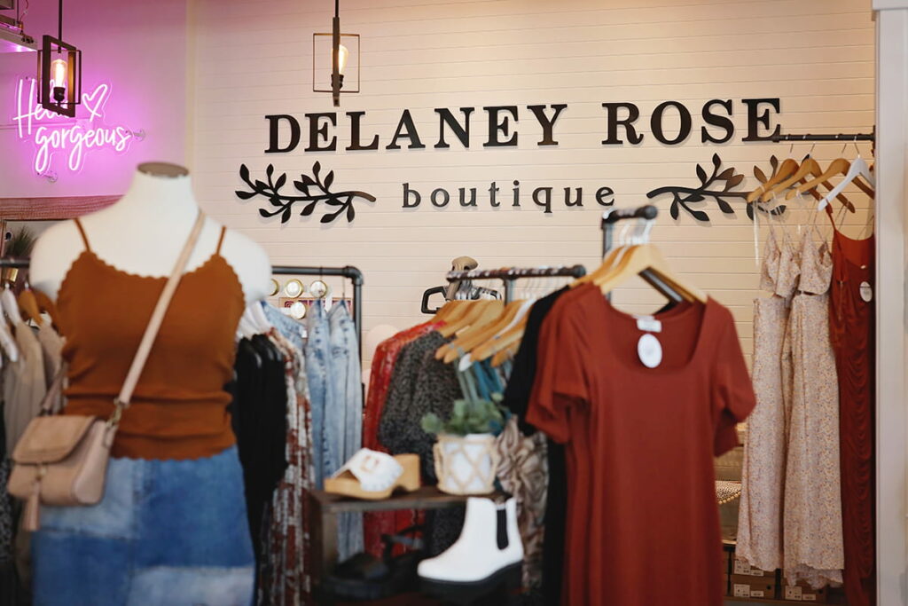 Trendy outfits displayed in the foreground with a sign for Delaney Rose Boutique visible on a wood paneled wall in the background