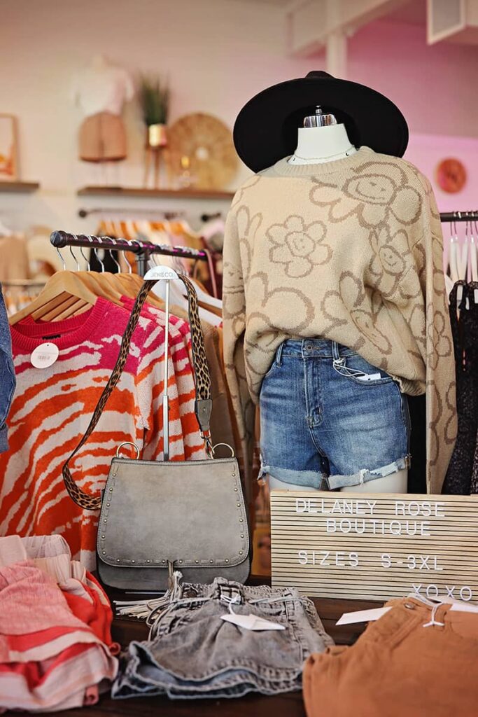A mannequin wearing jean shorts and a tan sweater covered in flowers with happy faces in the center is surrounded by clothing displayed neatly on racks. There is a sign in the foreground that reads "Delaney Rose Boutique | Sizes S - 3XL"