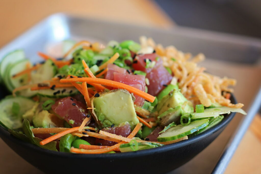 A black poké bowl loaded with edamame, sliced cucumber, carrot strips, chunks of tuna, and avocado. The bowl is topped with crispy noodles and black sesame seeds.