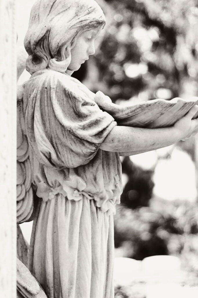 Side view of a statue of a young girl cradling a large bowl in her arms and looking down at it, the contents not visible to the viewer. The B&W photo shows beautiful old oak trees and Spanish moss in the background