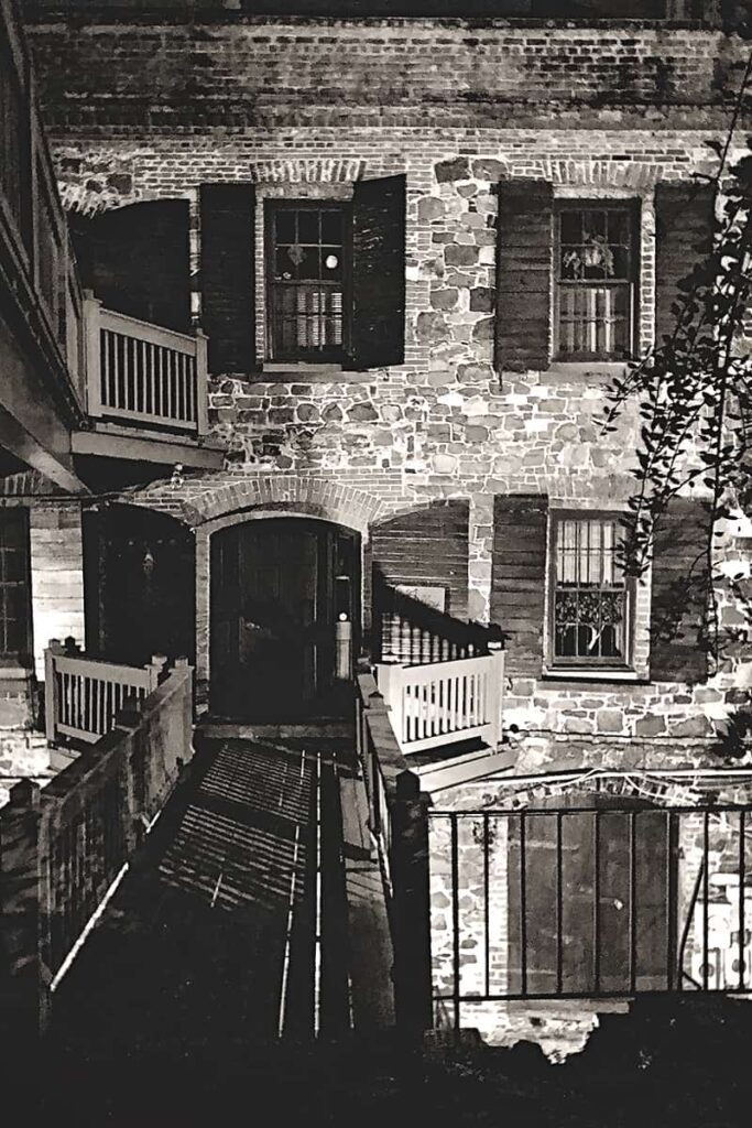 Spooky B&W shot of an old brick and cobblestone building in Savannah with a rickety wooden walkway leading to the door. The streetlights are causing wonky shadows on the building, making it appear haunted