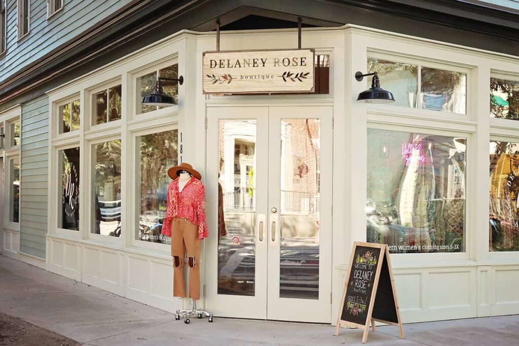 The front entrance to Delaney Rose Boutique in the Starland District with double glass doors and windows allowing a sneak peek into the shop. All of the trim is painted a pale, sage green, and a wooden sign with the store's logo painted in black hangs above the entrance