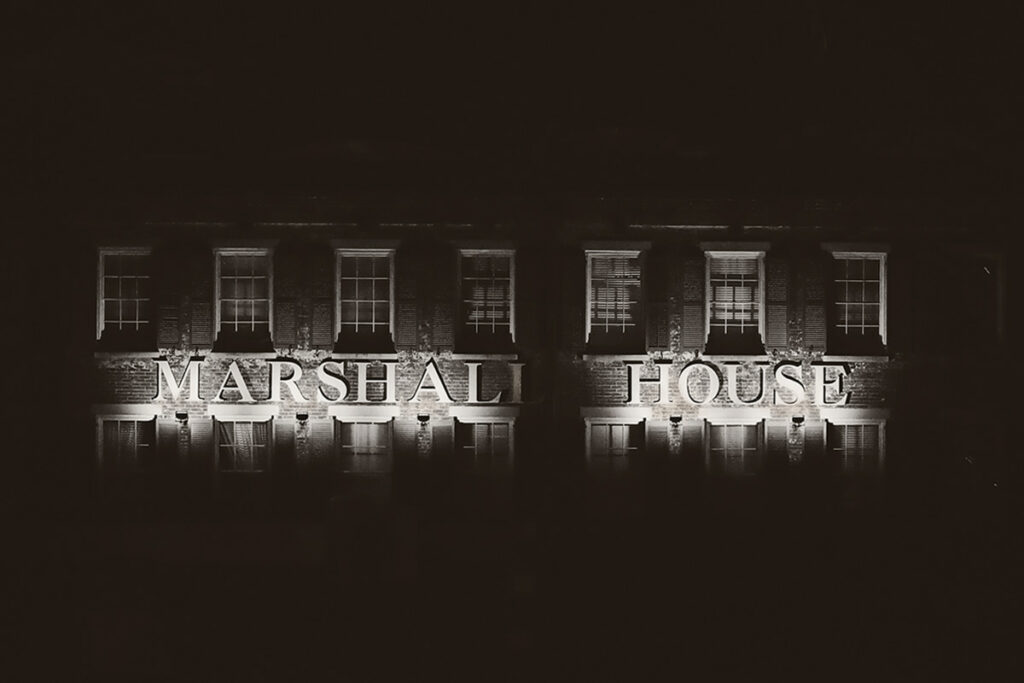 Large white letters spelling out the words "Marshall House" on the side of an old brick building. The building looks haunted because the photo is B&W and the sign is lit from below, casting eerie shadows over the facade of the building