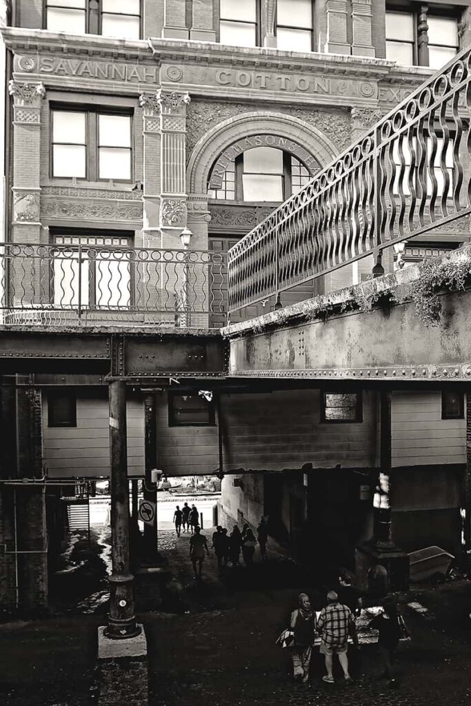 B&W image of the Historic Savannah Cotton Exchange Building with people walking along the Drayton Street Ramp beneath it