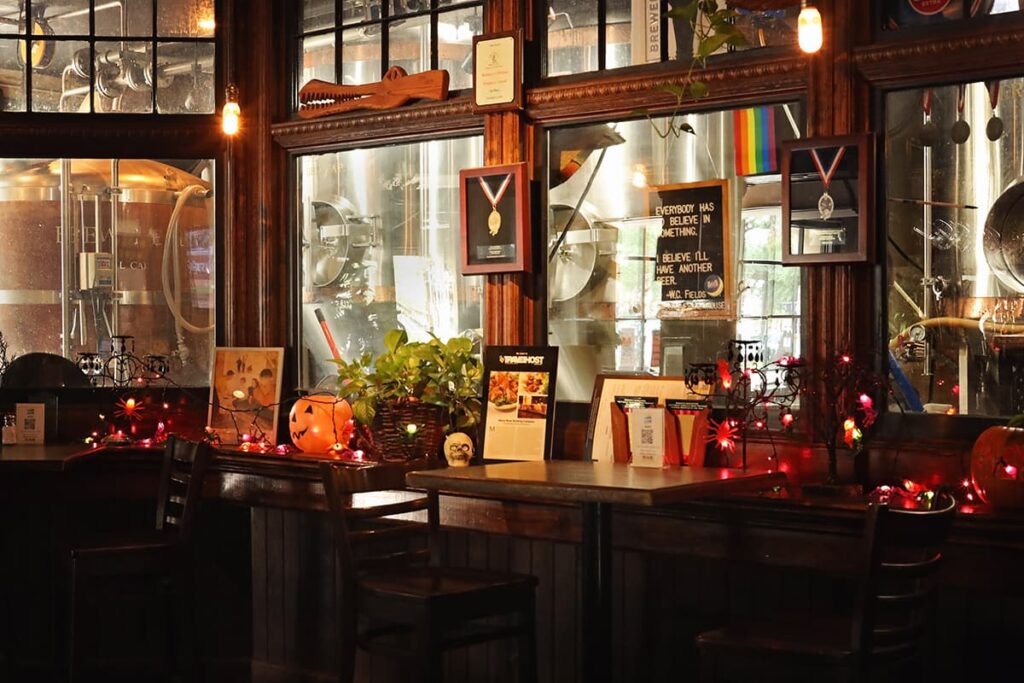 The interior of Moon River Brewing with hightop bar tables arranged next to windows where bar patrons can watch the brewing process as it occurs. The walls are trimmed in an elegant dark wood, Edison bulbs provide dim lighting, and Halloween decor is visible in a few areas