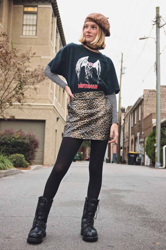 A thin blonde poses with her hand on her hip while standing in a lane in Savannah. She's wearing black leggings and boots, a leopard-print skirt, Mothman t-shirt layered over a B&W striped shirt, and a leopard-print cap
