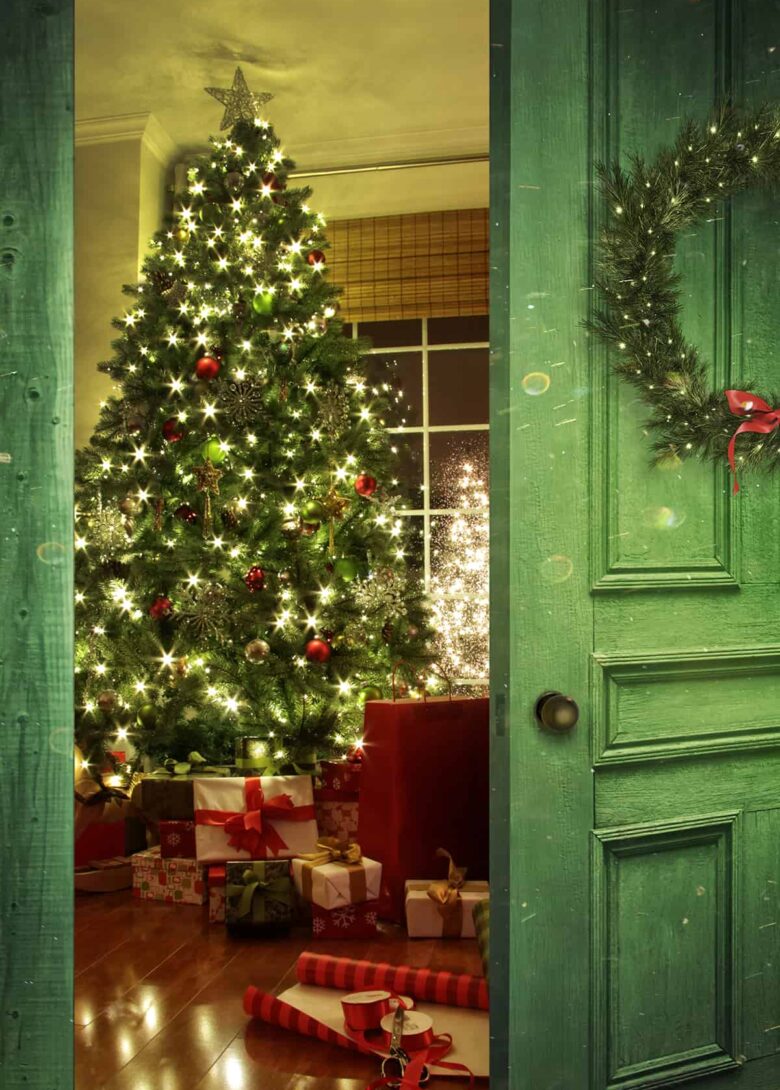 A cheery green door with a wreath on it is open to show the living room of a home filled with presents and a warmly-lit Christmas tree