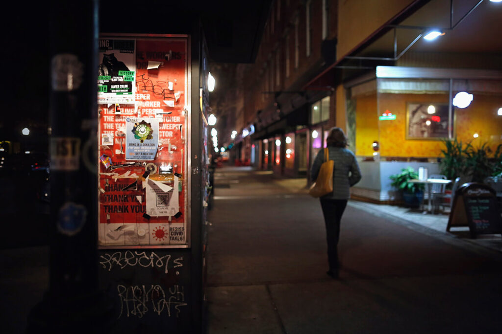 A woman walks down a dimly lit sidewalk on Broughton Street in Savannah. No one else is in sight, and she has just passed a sign covered in graffiti