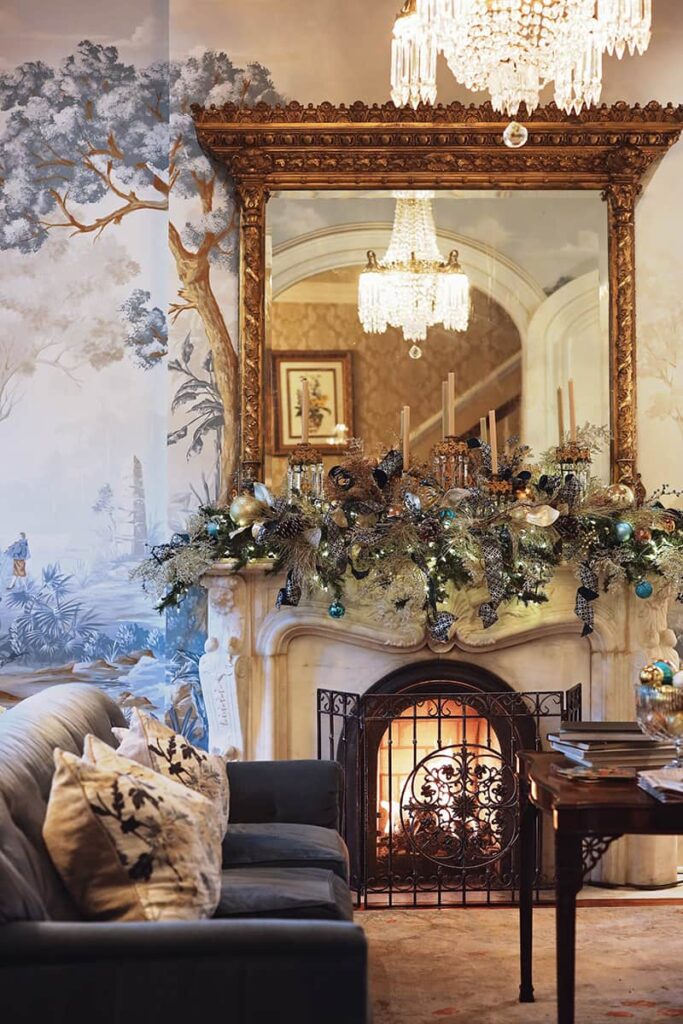 The parlor of Hamilton-Turner Inn shows a cozy seating area in front of a lit fireplace. The mantle is made from carved marble and a gilded mirror rests above it -- reflecting the room's chandelier. A swag of blue and brass ornaments on the mantle matches the designer wall mural