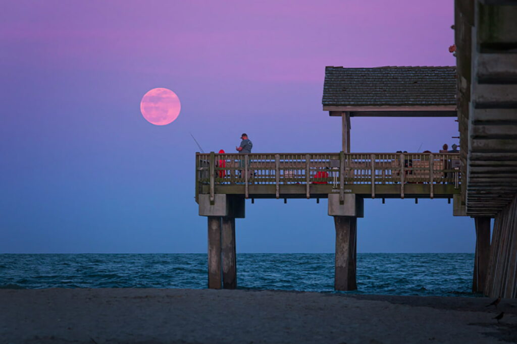 The Tybee Island pier at South Beach with a pink and lavender sunset visible over the water. A man and child are fishing off the pier with a pink full moon in the sky overhead