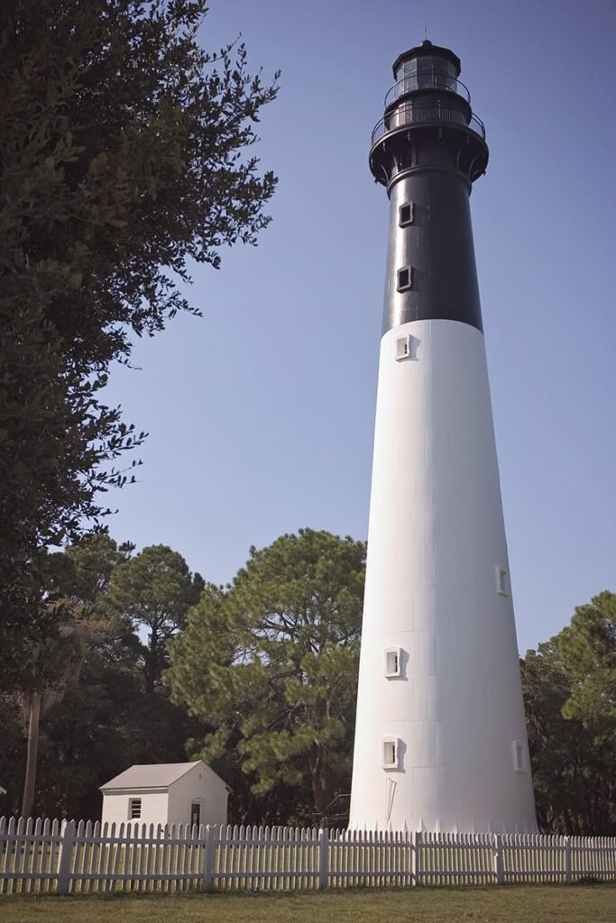 Full view of the B&W Hunting Island lighthouse with a white picket fence in the foreground and a small building and pine trees in the background