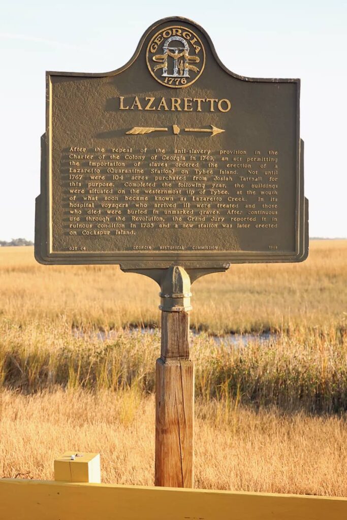 A Georgia Historical Society bronze marker with gold etchings indicates the definition of a Lazaretto as it applies to Tybee Island's history. Golden marsh reeds and a small amount of water at low tide are visible in the background
