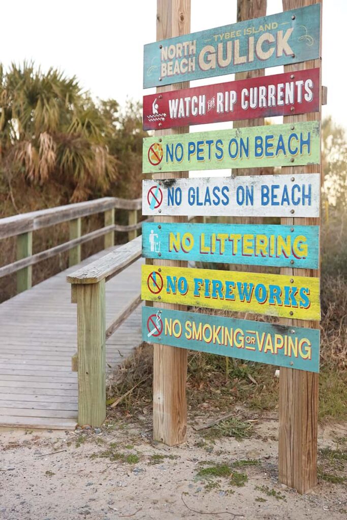 A boardwalk lined with palm trees and a colorful sign indicating it's the Tybee Island North Beach Gulick Street entrance. Each board is painted in a different color and lists a separate beach rule, such as "Watch the Rip Currents" "No pets on the beach" "No fireworks", etc.