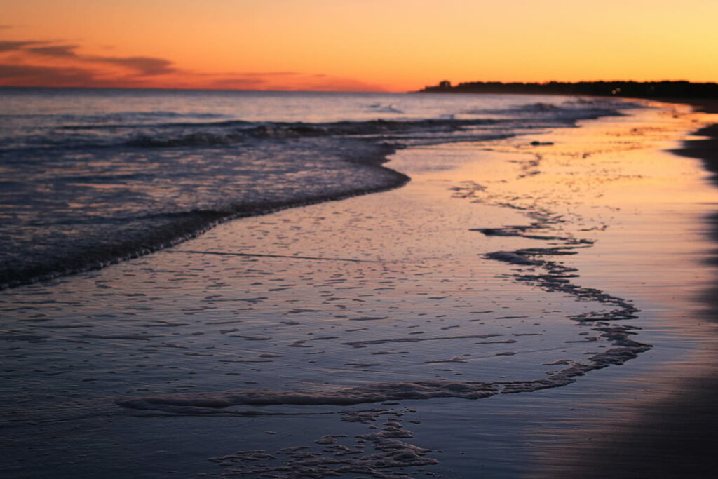 Soft waves lap against the sand on Folly Field Beach in Hilton Head as an orange and purple sunset lights the sky