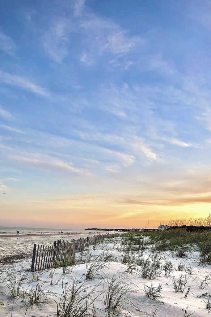 Hilton Head is one of the prettiest beaches near Savannah GA, with white and wooden fences to protect the dunes. Blue skies with trails of white clouds are visible in the distance and fade into a pale peach and orange sunset