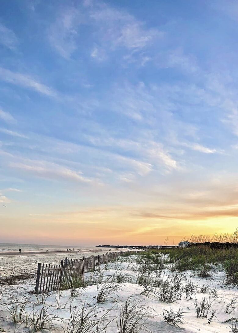 Hilton Head is one of the prettiest beaches near Savannah GA, with white and wooden fences to protect the dunes. Blue skies with trails of white clouds are visible in the distance and fade into a pale peach and orange sunset
