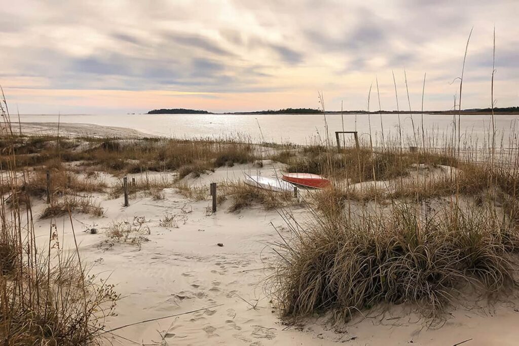 A cloudy fall day on Tybee's Back River Beach shows marsh grasses fading to brown and kayaks stored between the dunes. A sunset fades to pink and Little Tybee Island is visible in the distance