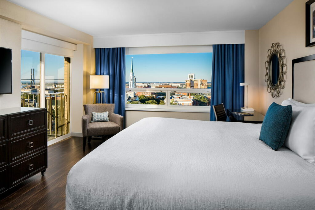 Hotel room at The DeSoto Savannah showing a king-sized bed with a white bedspread. Sliding glass doors lead out to a balcony with beautiful rooftop views of Savannah's Historic District