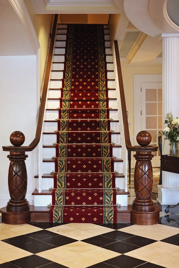 A staircase inside The Marshall House features beautiful hand-carved railings designed to look like pineapples. The treads are painted white and a burgundy and gold runner extends the entire length of the stairs. The floor of the hotel's entrance is an elegant B&W large diamond-checkered pattern