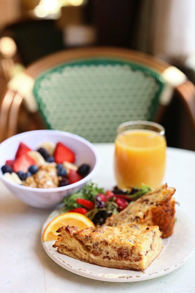 Two hearty slices of quiche next to a colorful salad on a plate in the foreground, with a bowl of granola in the background alongside a full glass of orange juice