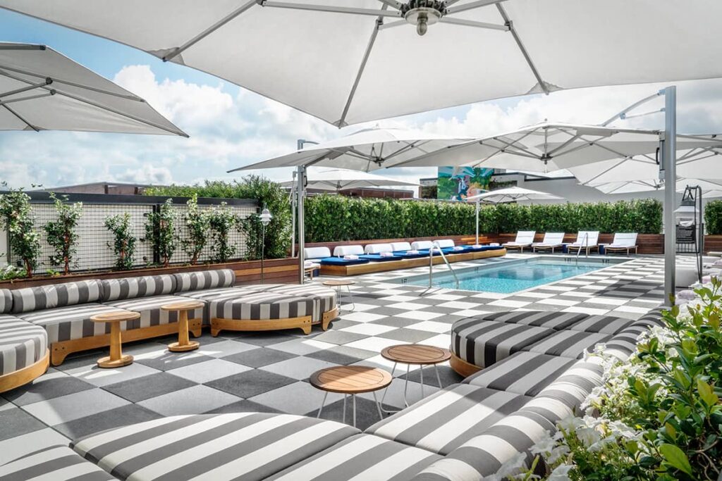 The rooftop pool deck at Perry Lane Hotel features ample seating upholstered in grey and white stripes, teak tables, poolside lounging zones, and a few white tented areas to provide shade for those who avoid full sun