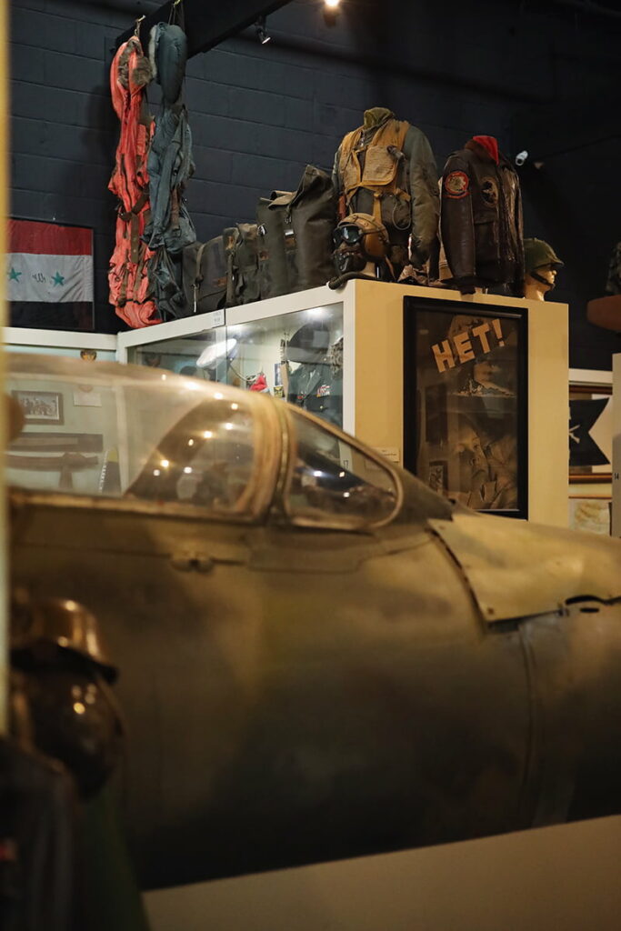 The front portion of a military plane is on display in the foreground of Webb Military Museum while a glass case full of uniforms and posters is visible in the background