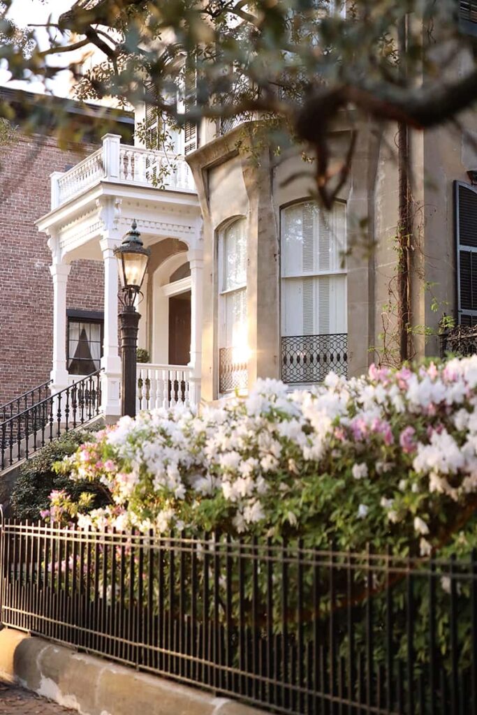 Stately home in Savannah with an intricately detailed white front porch, a gas lantern near the door, and hundreds of pink and white azaleas growing over a wrought iron fence