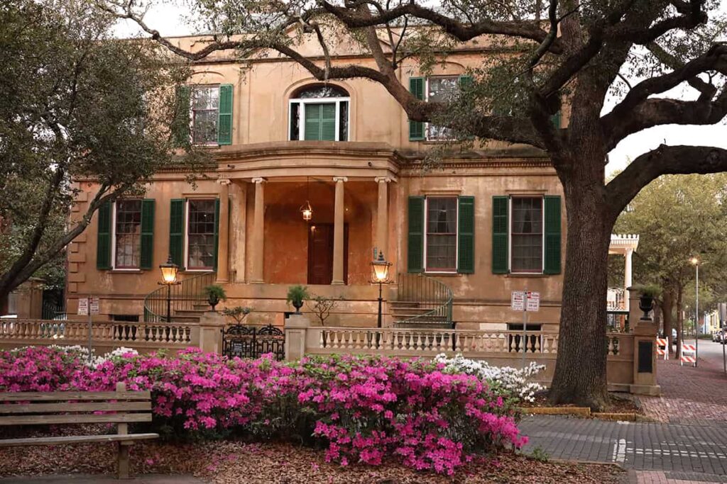 Front façade of the Owens Thomas House, with orange toned walls and green shutters surrounding the windows. The foreground is filled with pink and white azalea bushes and a park bench