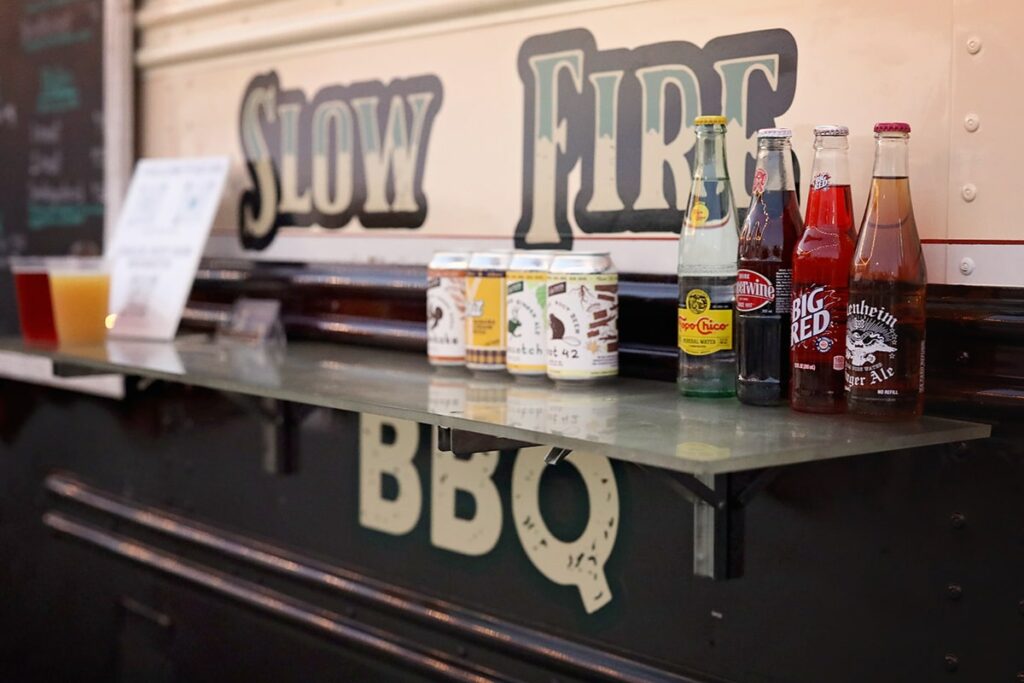 The Slow Fire BBQ logo on the side of their food truck, with text written in green and white with a black outline. A selection of drinks sits on a shelf in front of the sign