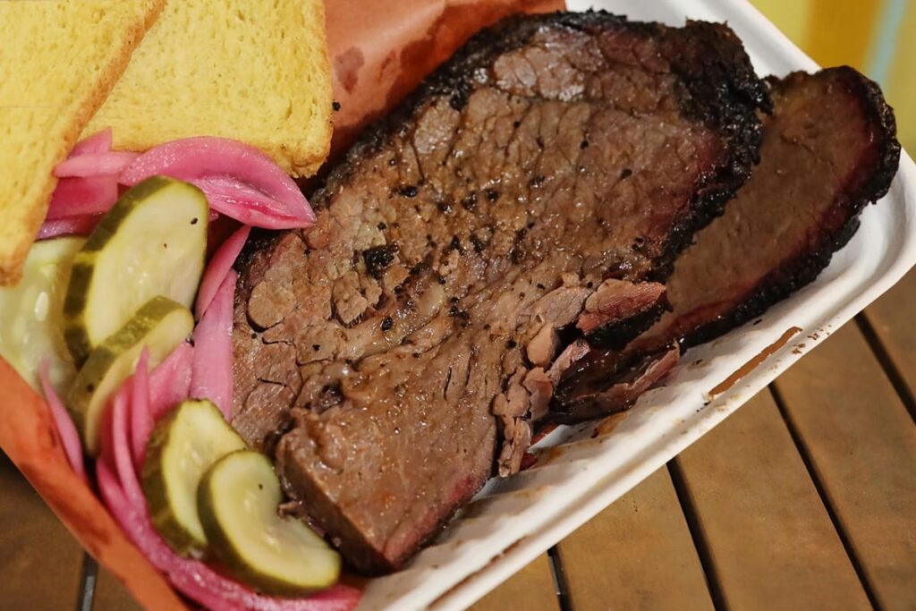 Thick slice of tender brisket in a styrofoam container surrounded by bread, pickles, and onion