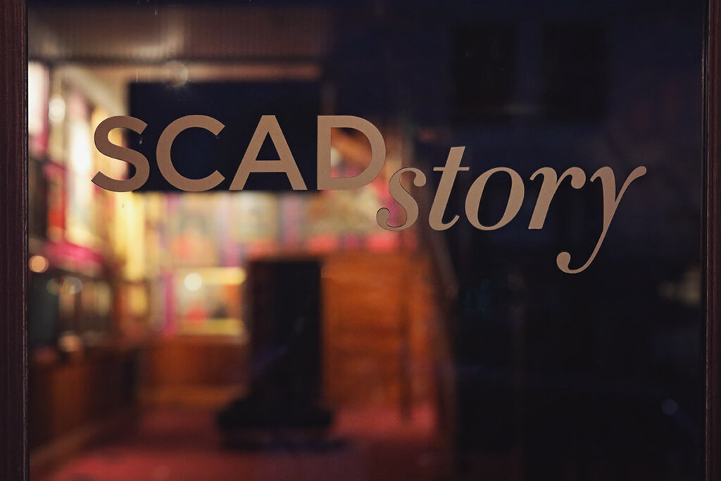 The SCADstory logo on the glass door entrance to the museum, with a colorful plaid wall covered in art visible in the background