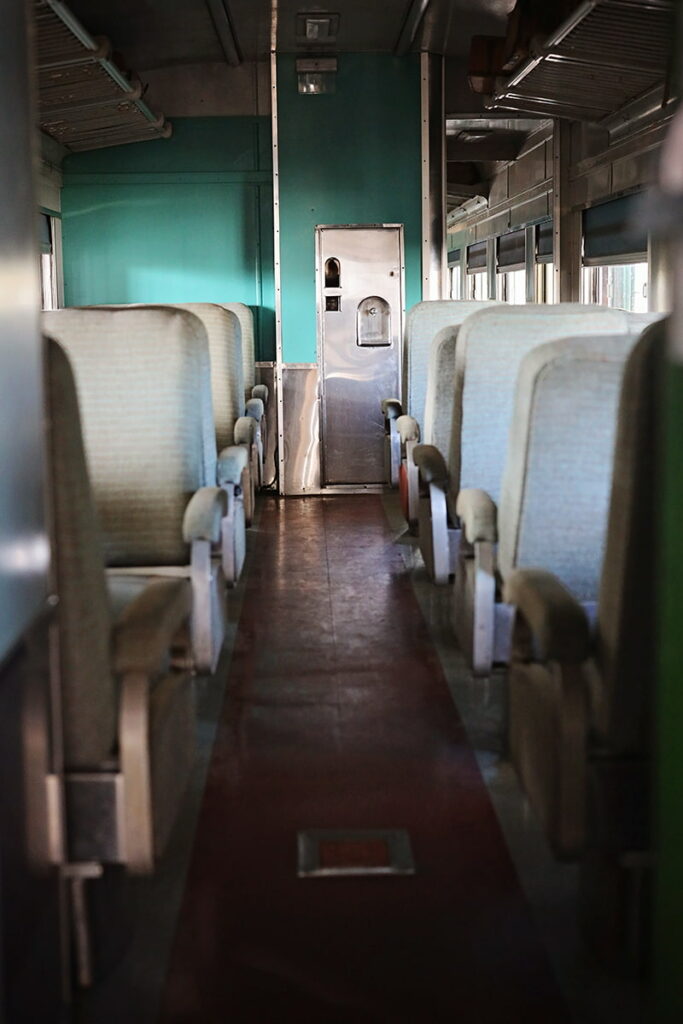 A scuffed dark red floor leads visitors down the interior of a railcar, past passenger seating and towards a teal green colored wall with a small metal door