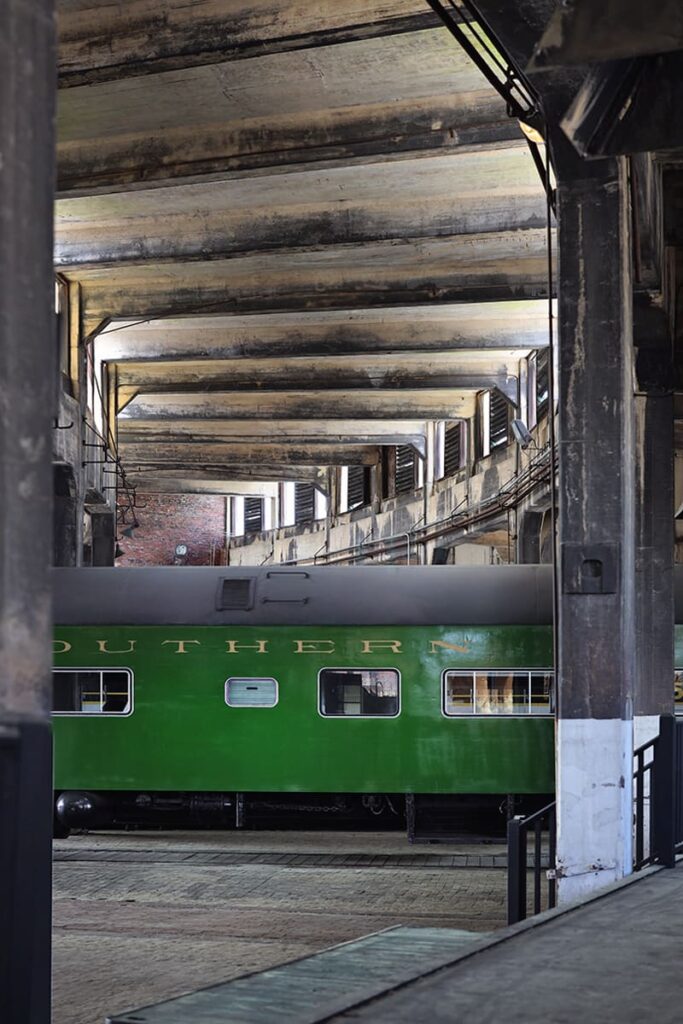 Rusted steel beams along the ceiling of the Roundhouse at the Georgia State Railroad Museum contrast sharply against a bright green railcar with the word "SOUTHERN" imprinted across the side in yellow text